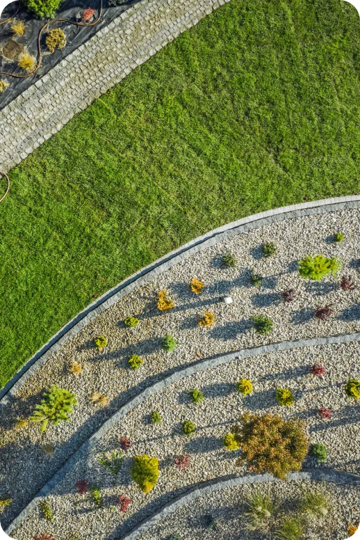 Aerial view of a landscaped garden with curved pathways. On the left is a lush green lawn, and on the right, neatly arranged plants on gravel.
