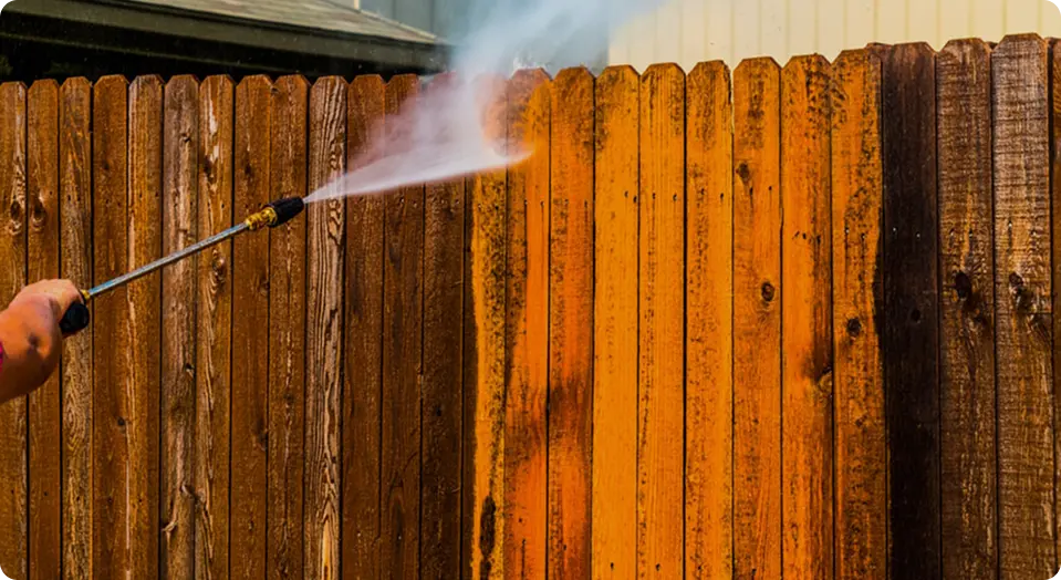 A person power washing a wooden fence, revealing vibrant orange beneath the dirt. The contrast highlights the cleaning process. Energetic and transformative tone.