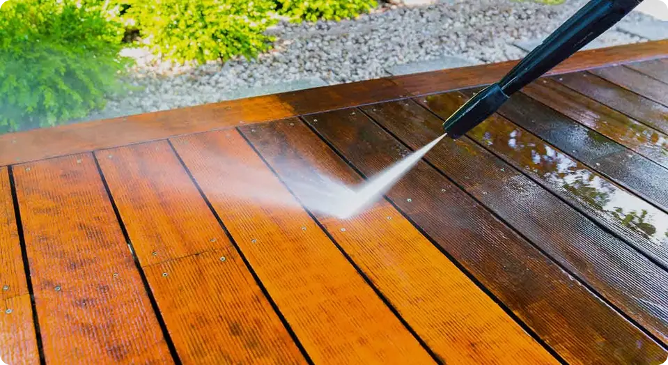 A pressure washer cleaning a wooden deck, showing a clear contrast between the clean and dirty areas. The background has green bushes and gravel.
