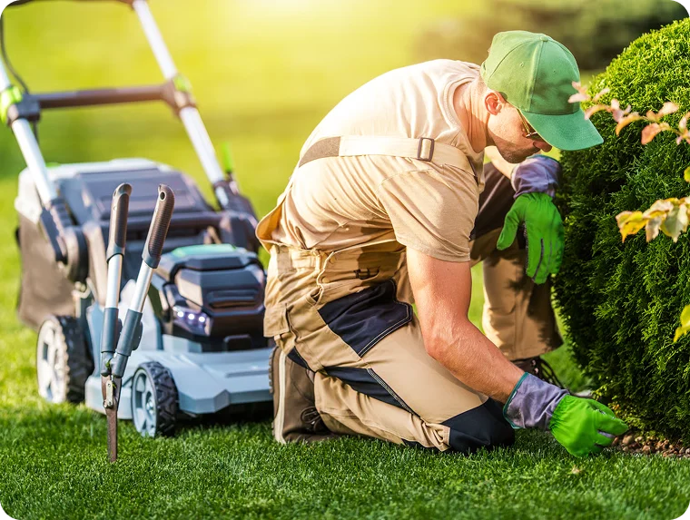 Landscaper kneeling beside a lawn mower to tidy a hedge for absentee landlord lawn care in Vancouver